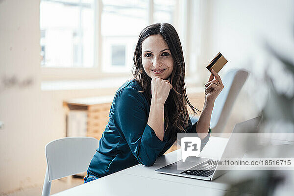 Smiling businesswoman holding credit card sitting at table in home office