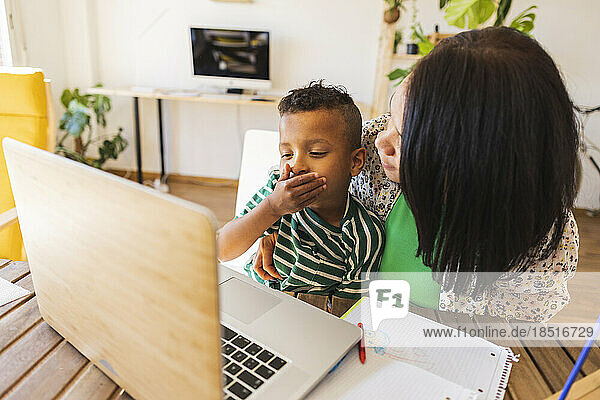 Boy covering mouth with mother watching laptop at home