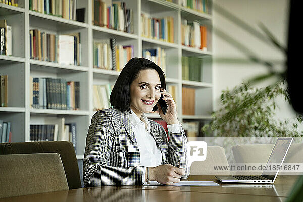 Smiling businesswoman talking on smart phone sitting at desk in office