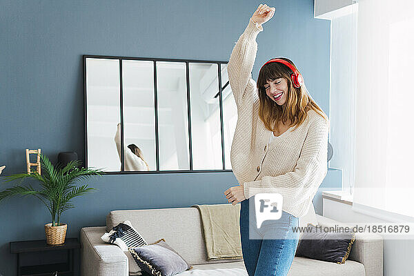 Carefree woman wearing headphones dancing with hand raised in living room at home