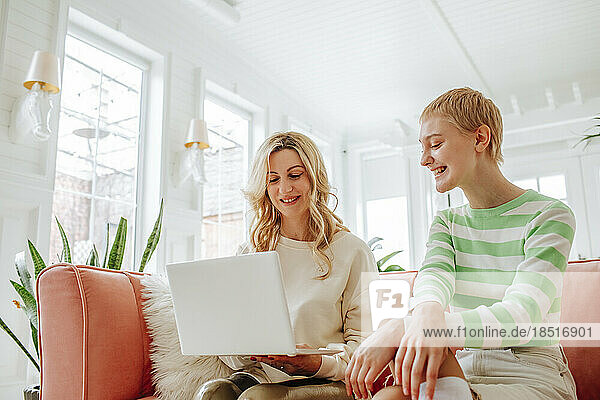 Smiling woman sharing laptop with friend sitting on sofa in living room