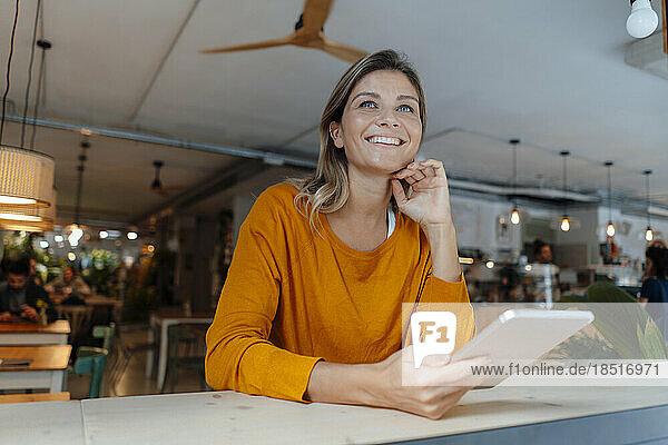 Happy woman holding tablet PC sitting with hand on chin in cafe