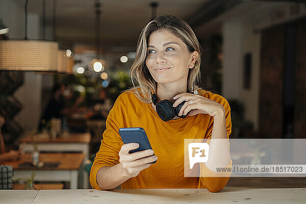Nachdenklich lächelnde Frau mit Kopfhörern und Mobiltelefon sitzt im Café