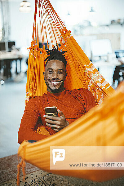 Happy young businessman relaxing in hammock with smart phone in office
