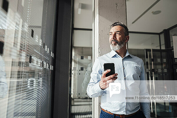 Thoughtful businessman standing with smart phone in office