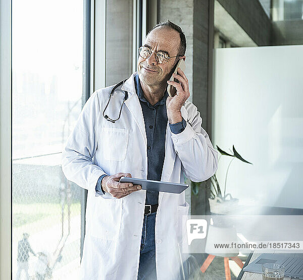 Mature doctor with tablet PC talking on smart phone by window