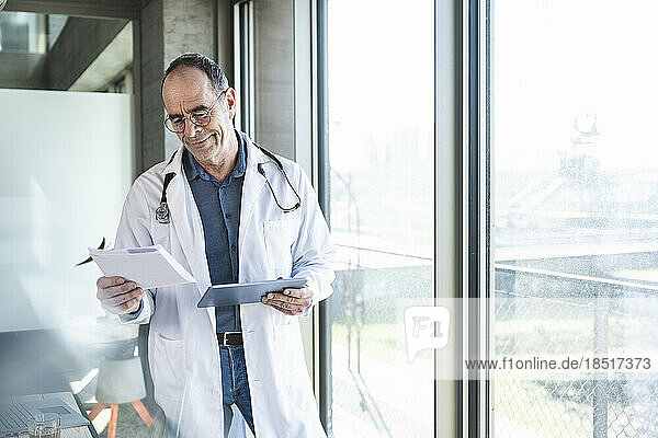 Smiling mature doctor reading prescription and holding tablet PC by window
