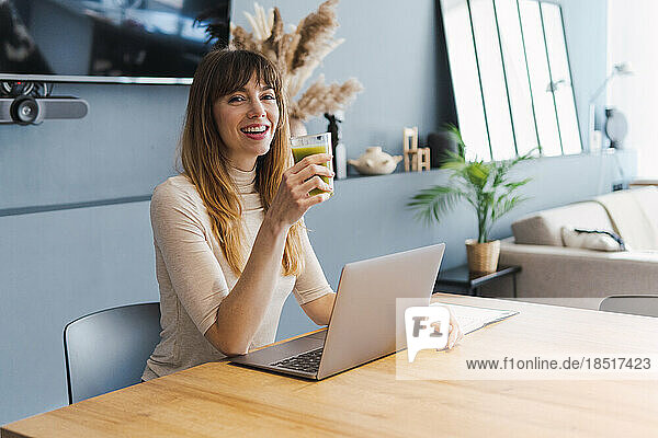 Glücklicher Freiberufler hält Detox-Saft in der Hand und sitzt mit Laptop am Schreibtisch im Heimbüro