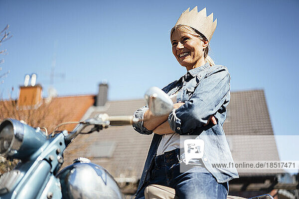 Happy mature woman wearing crown standing with arms crossed on motorcyle