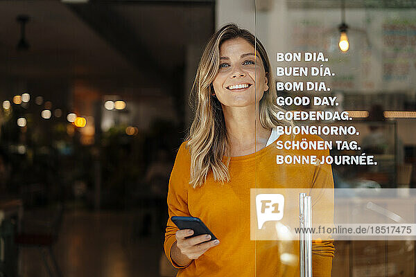Happy woman with blond hair holding smart phone standing at door