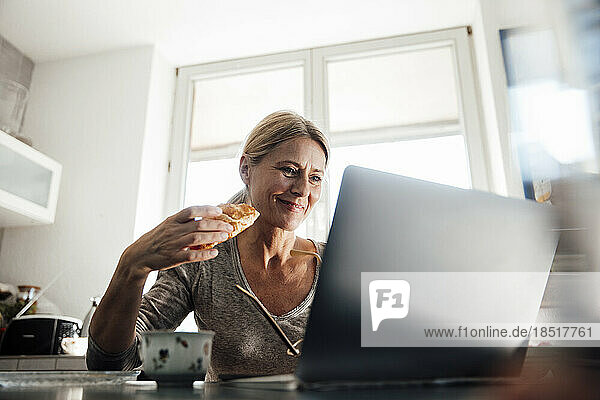 Smiling freelancer with croissant working on laptop at home office