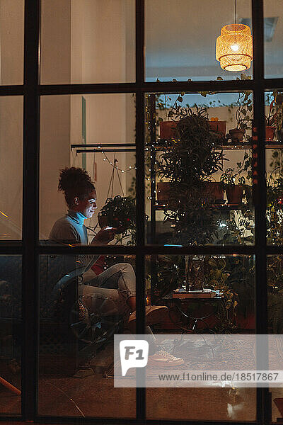 Geschäftsfrau benutzt Mobiltelefon nachts auf Sofa  gesehen durch Glas