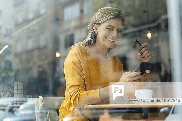 Happy woman using smart phone with credit card in cafe seen through glass