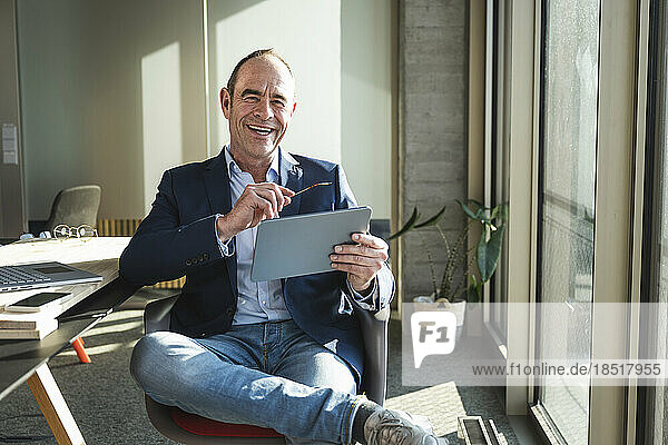 Happy mature businessman with tablet PC sitting in office