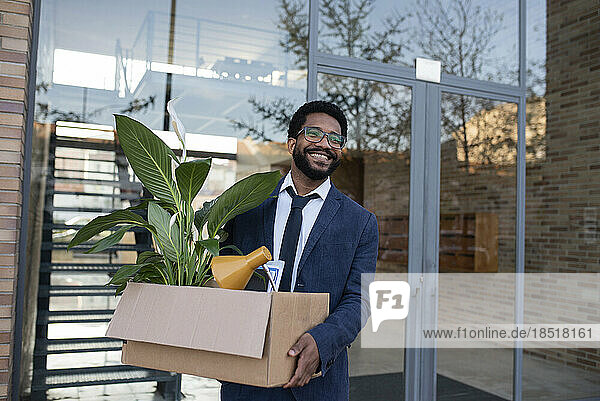 Lächelnder Geschäftsmann verlässt Bürogebäude mit Karton