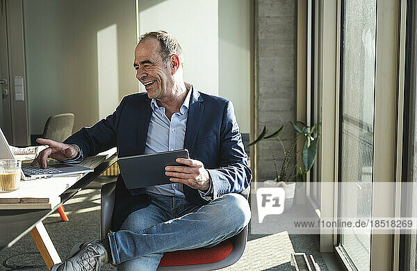 Happy businessman using laptop and holding tablet PC in office