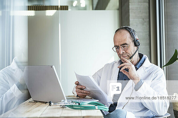 Doctor wearing headset reading document sitting with laptop in office