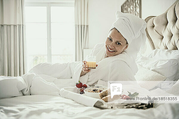 Happy woman holding juice glass by breakfast tray on bed at home