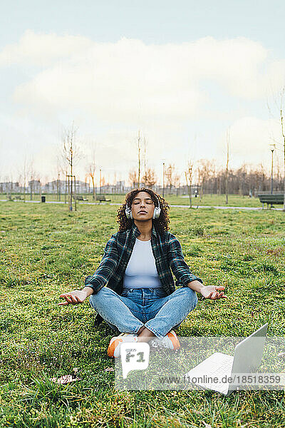 Young woman wearing wireless headphones meditating in park at sunset