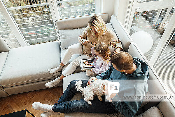 Daughter looking at photos with parents and dog sitting on sofa at home