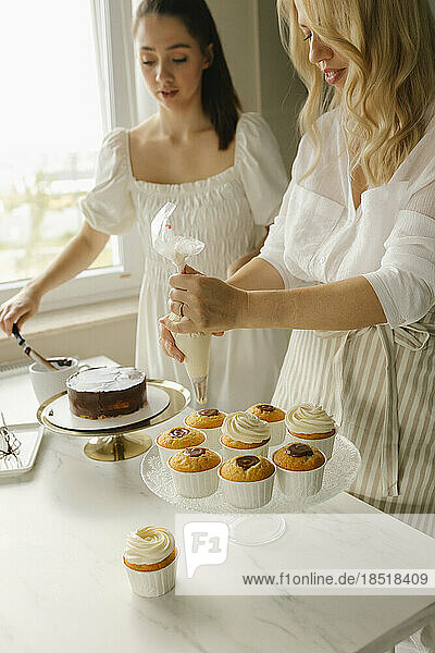 Woman with friend preparing cake and decorating cupcakes at home