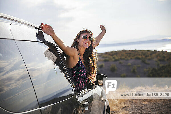 Happy young woman with arms raised leaning out of car window