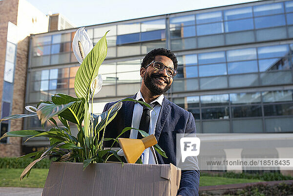 Lächelnder Geschäftsmann trägt Karton mit Büromaterial vor Bürogebäude