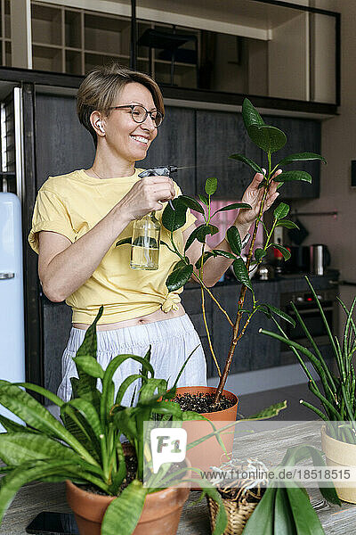Smiling woman spraying water on plant leaves at home