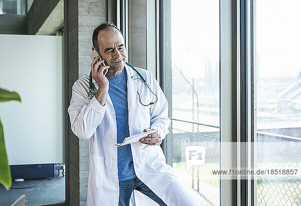 Smiling mature doctor talking on smart phone standing by window