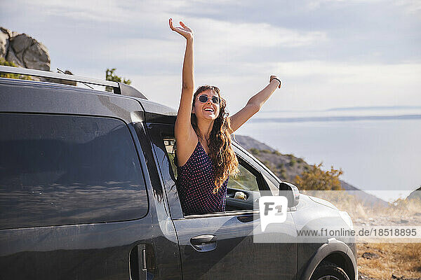 Cheerful young woman with arms raised leaning out of car window