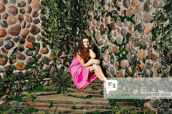 Eine erwachsene Frau sitzt in ihrem hübschen Sommerkleid  mit gewelltem Haar und einem charmanten Lächeln auf einer Steinplatte nahe der Mauer. Sie sieht entspannt und zufrieden aus und genießt das Wochenende im Freien