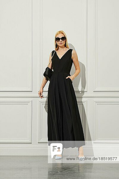 Stilvolle blonde Geschäftsfrau in schwarzem Kleid und Sonnenbrille hält Handtasche auf ihrer bloßen Schulter