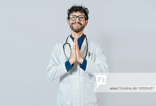 Lächelnder Arzt  der seine Handflächen auf einem isolierten Hintergrund zusammenlegt. Schöner Arzt mit Händen zusammen isoliert. Junger Arzt mit zusammengelegten Handflächen
