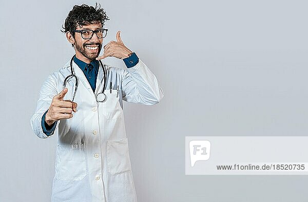 Junger Arzt macht Anruf Geste isoliert. Lächelnder Arzt macht Anruf Geste und zeigt auf die Kamera
