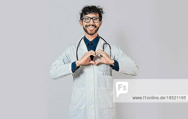 Junger Arzt macht Herzform isoliert. Hübscher Arzt macht Herz Geste mit den Händen. Glücklicher Arzt zeigt mit dem Finger nach unten