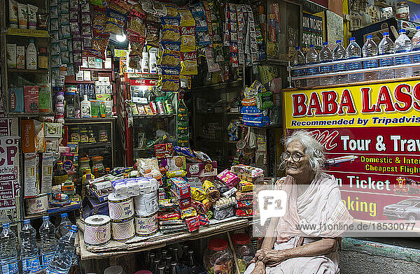 Eine ältere Ladenbesitzerin sitzt in ihrem Laden voller verschiedener Waren; Varanasi  Indien