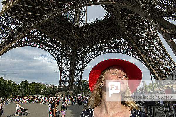 Junge Frau mit rotem Hut steht unter dem Eiffelturm und sonnt sich im Sonnenlicht in Paris  Frankreich; Paris  Frankreich