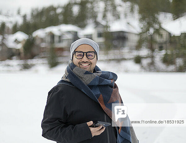 Porträt eines jungen Mannes im Freien an einem Wintertag  der eine Brille  einen Mantel und eine Strickmütze trägt und ein Smartphone in der Hand hält; British Columbia  Kanada