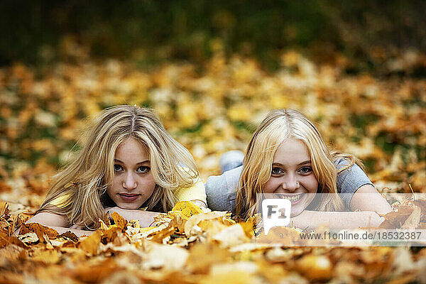 Portrait of two teenage girls laying in the leaves and posing for the camera in a city park on a warm fall afternoon; St. Albert  Alberta  Canada.