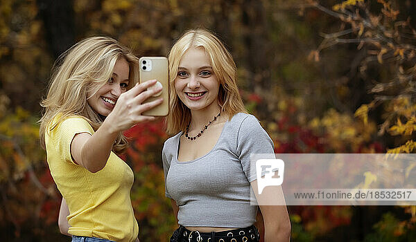 Two teenage girls taking self-portraits with a smart phone while spending time together in a city park on a warm fall day; St. Albert  Alberta  Canada.