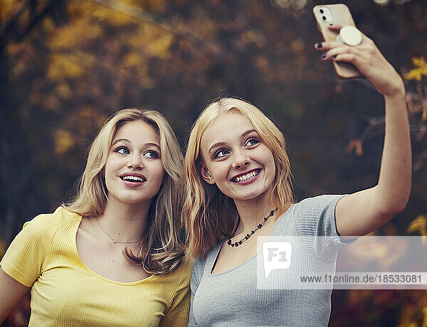 Two teenage girls taking self-portraits with a smart phone while spending time together in a city park on a warm fall day; St. Albert  Alberta  Canada.