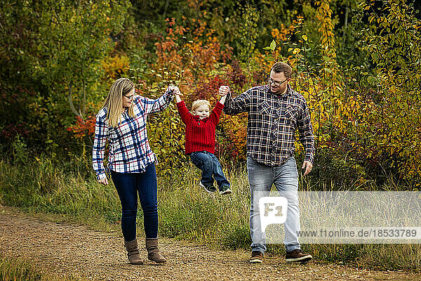 Mutter und Vater halten sich mit ihrem kleinen Sohn an den Händen  während sie im Herbst einen Weg in einem Stadtpark entlanggehen; Edmonton  Alberta  Kanada