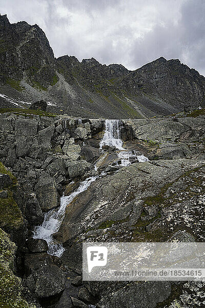 Glacial waterfall over rocky terrain at Archangel Hatcher Pass with the granite mountain peaks in the background under a grey  cloudy sky near Independence Mine; Palmer  Alaska  United States of America