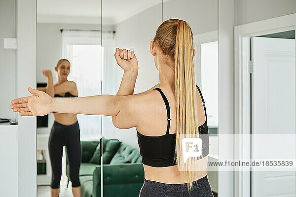 Young woman in sportswear stretches arms in her living room in front of a mirror  back view