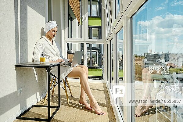 Barfüßige Frau im Bademantel sitzt auf dem Balkon ihrer neuen Hochhauswohnung und benutzt einen Laptop