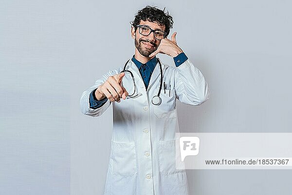 Ein lächelnder Arzt macht eine Geste und zeigt auf die Kamera. Junger Arzt macht Anruf Geste isoliert