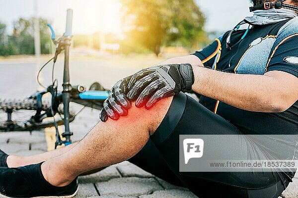 Concept of cyclist with knee injury outdoors. Cyclist with knee pain outdoors. Male cyclist sitting on the pavement with knee pain