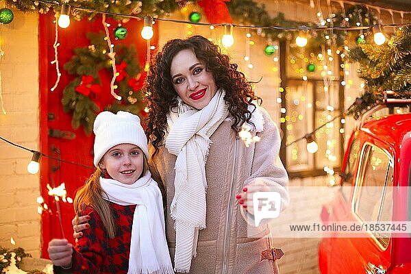 Fröhliche Feiertage. Mutter und kleine Tochter halten Wunderkerzen und lächeln in die Kamera  während sie im Freien vor einem für Weihnachten und Neujahr dekorierten Haus stehen. Mutter und Kind feiern Weihnachten im Freien
