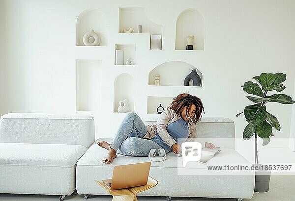 Foto von hübscher glücklicher junger afrikanischer Frau  die zu Hause auf dem Sofa sitzt und eine Zeitschrift oder ein Buch liest
