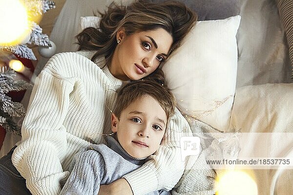 Cozy christmas portrait of young family mother and little son smiling at camera while relaxing together on sofa near xmas tree at home  mom and kid enjoying winter holidays together. View from above